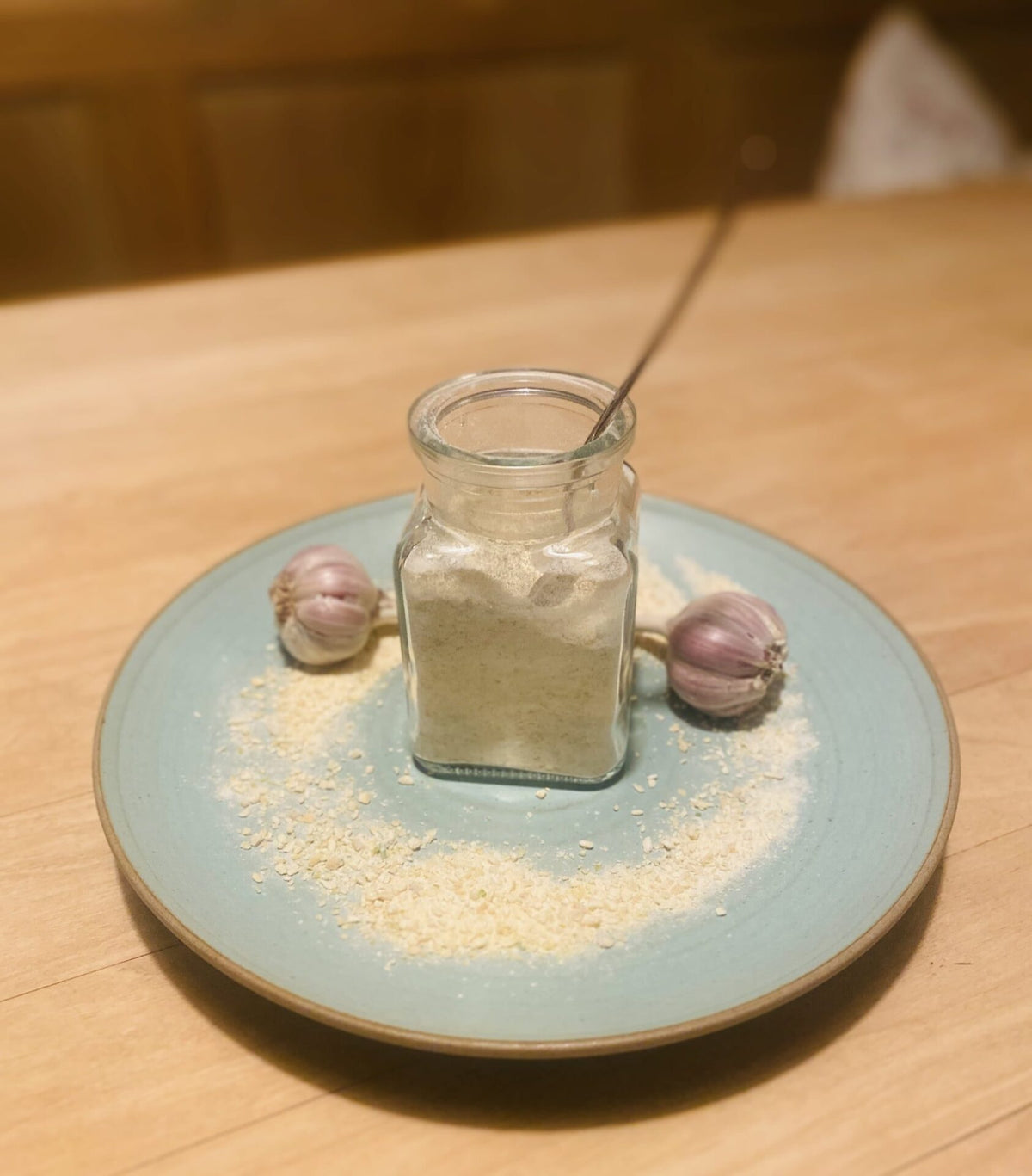 Glas mit Löffelglück Knoblauch-Konzentrat auf hellblauem Teller, umgeben von Granulat und zwei kleinen Knollen
