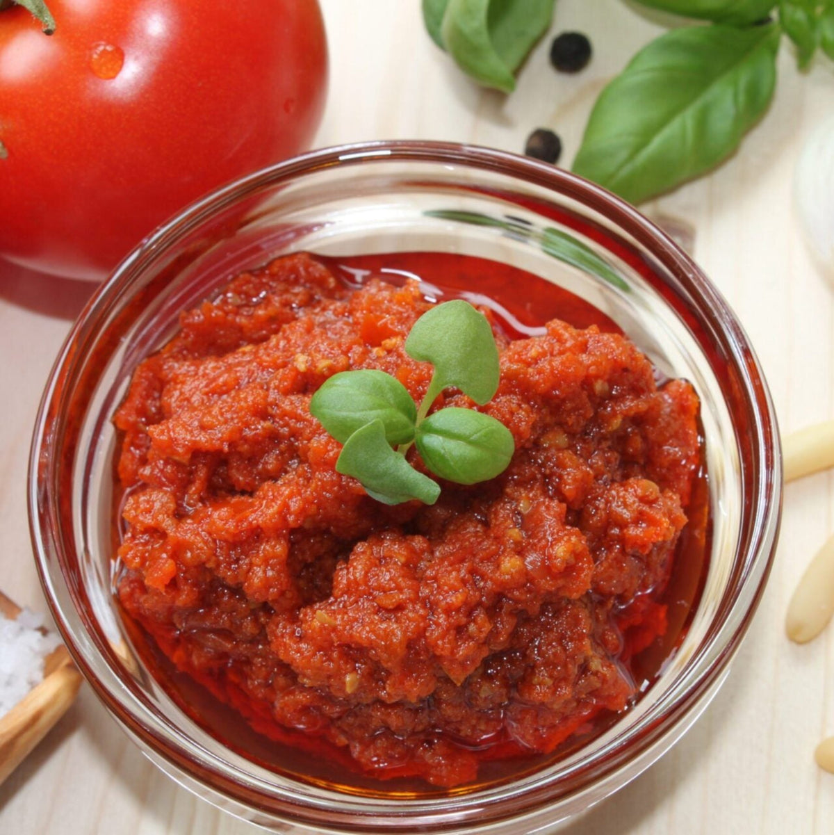 Veganes Knobi-Pesto in einer Glasschale mit Basilikum, daneben Tomate und Pinienkerne