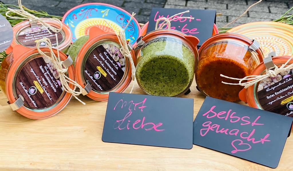 Weck-Gläser mit grünem und rotem Knobi-Pesto auf einem Holztisch, davor Tafeln mit Aufschrift mit Liebe selbst gemacht