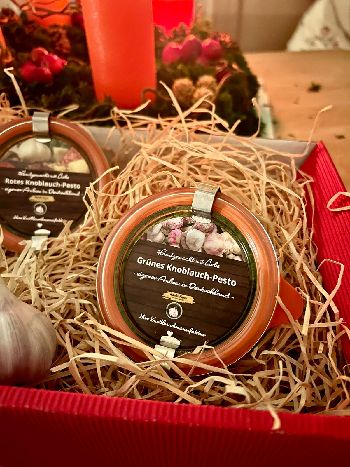 Rote Geschenkbox mit Holzwolle und Weck-Gläsern mit rotem und grünem Knoblauch-Pesto, dahinter ein Adventsgesteck