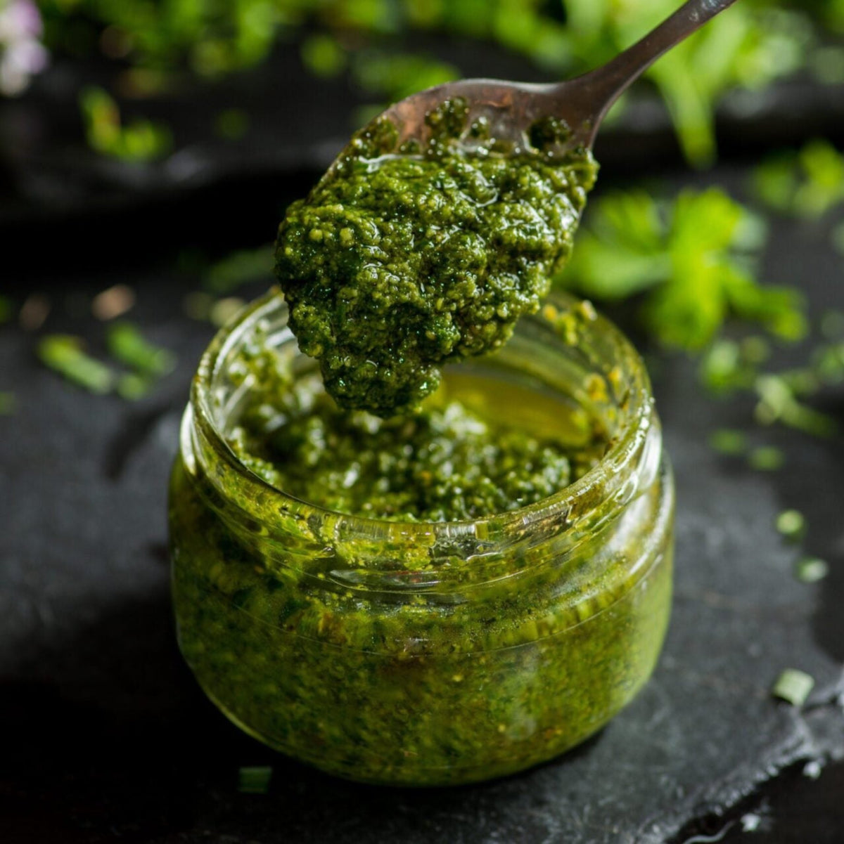 Löffel hebt Grünes Knobi-Pesto aus einem Glas, umgeben von frischen Kräutern auf dunklem Untergrund