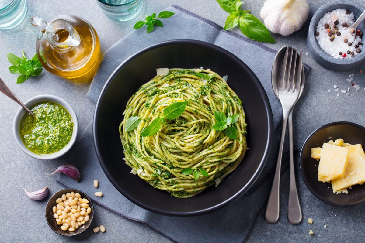 Teller Spaghetti mit grünem Pesto und Basilikum, daneben Pinienkerne, Parmesan und Öl