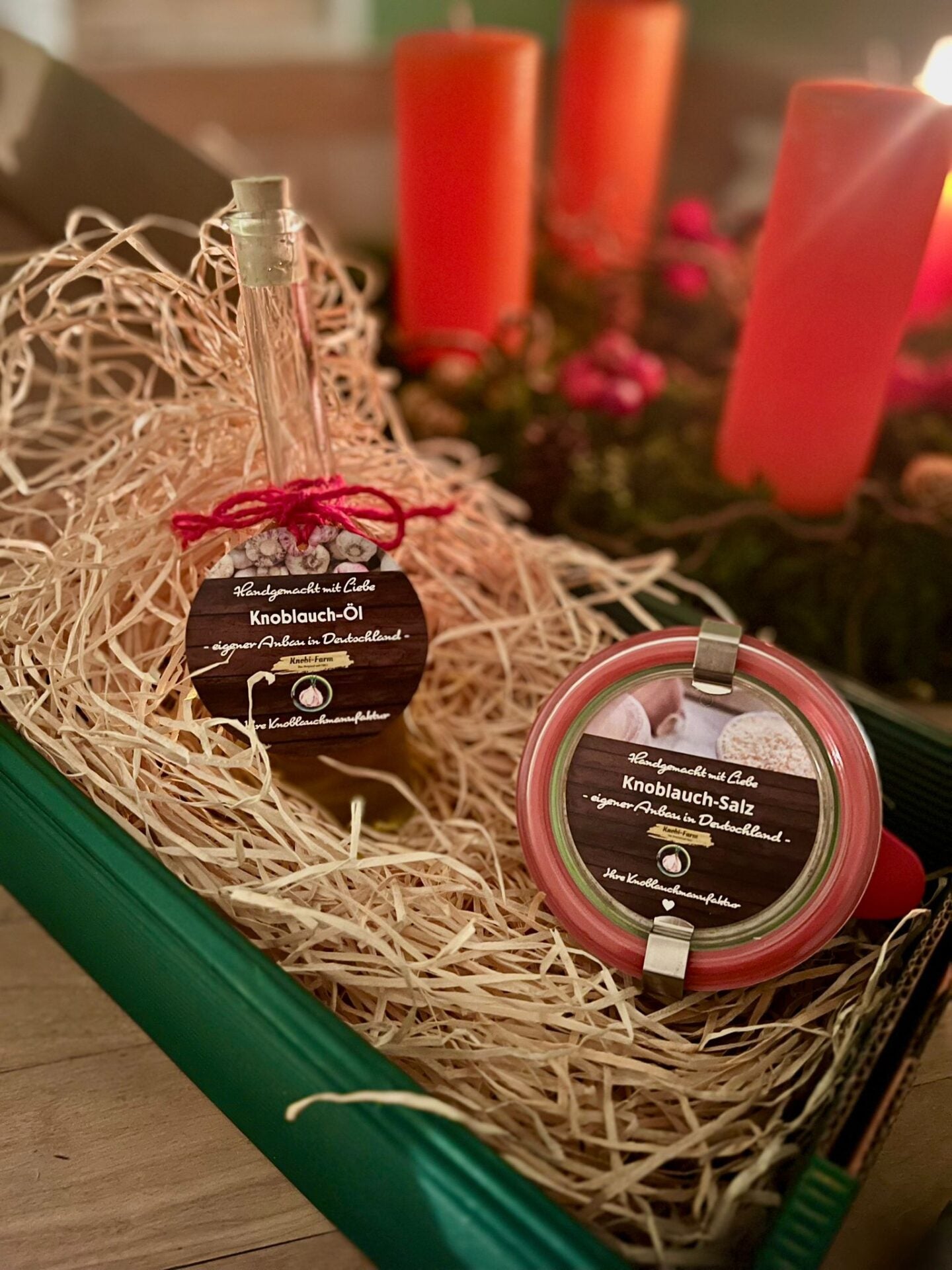 Knobi-Genuß Geschenk-Set: Flasche Knoblauch-Öl und Glas Knoblauch-Salz in Holzwolle, dahinter rote Kerzen