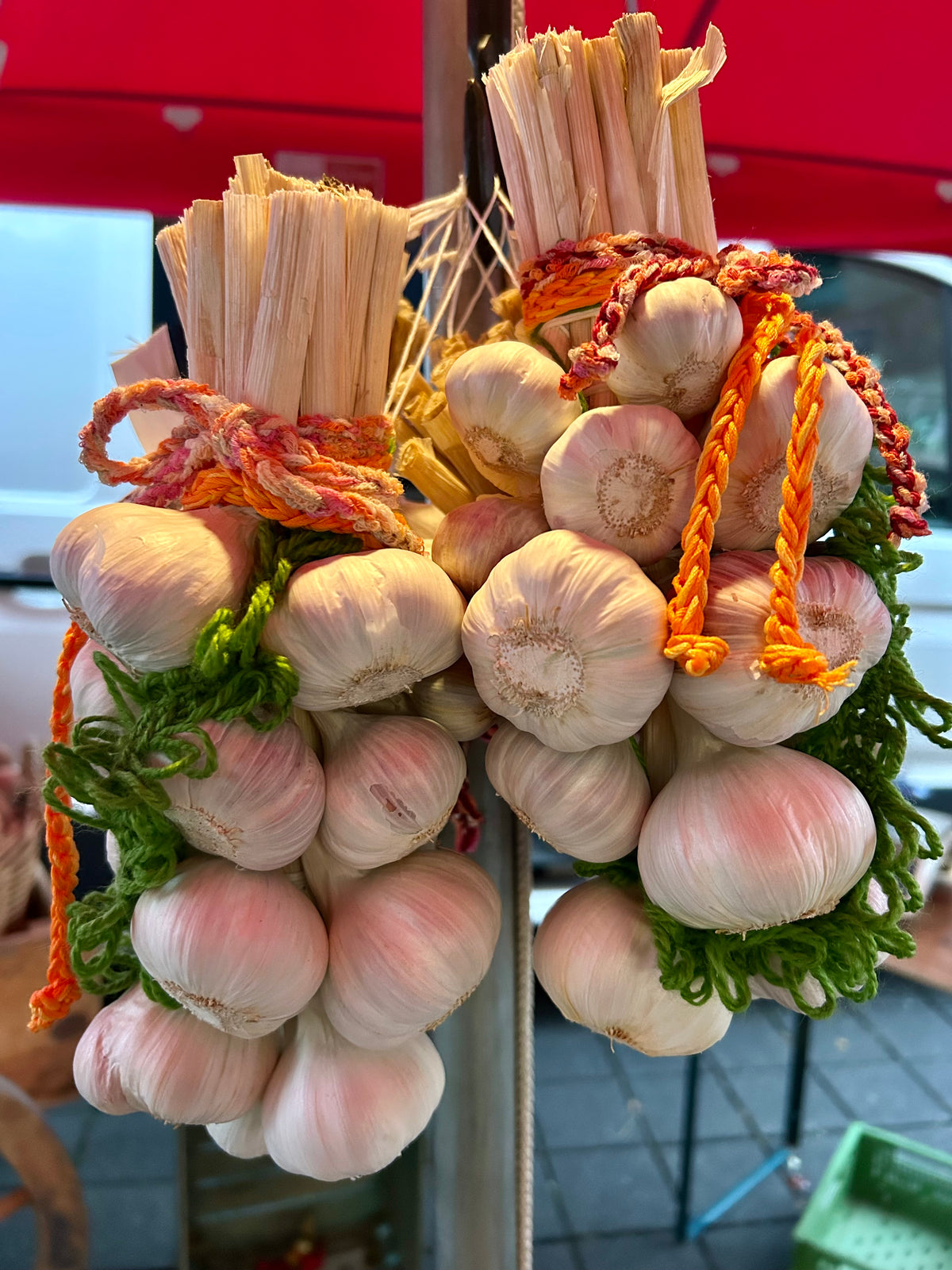 Zwei handgebundene Knobi-Zöpfe mit vielen Knoblauchknollen hängen an einem Marktstand