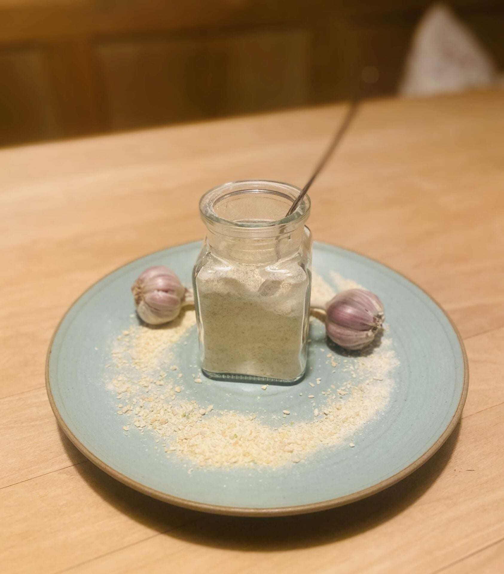 Glas mit Löffelglück Knoblauch-Konzentrat auf hellblauem Teller, umgeben von Granulat und zwei kleinen Knollen