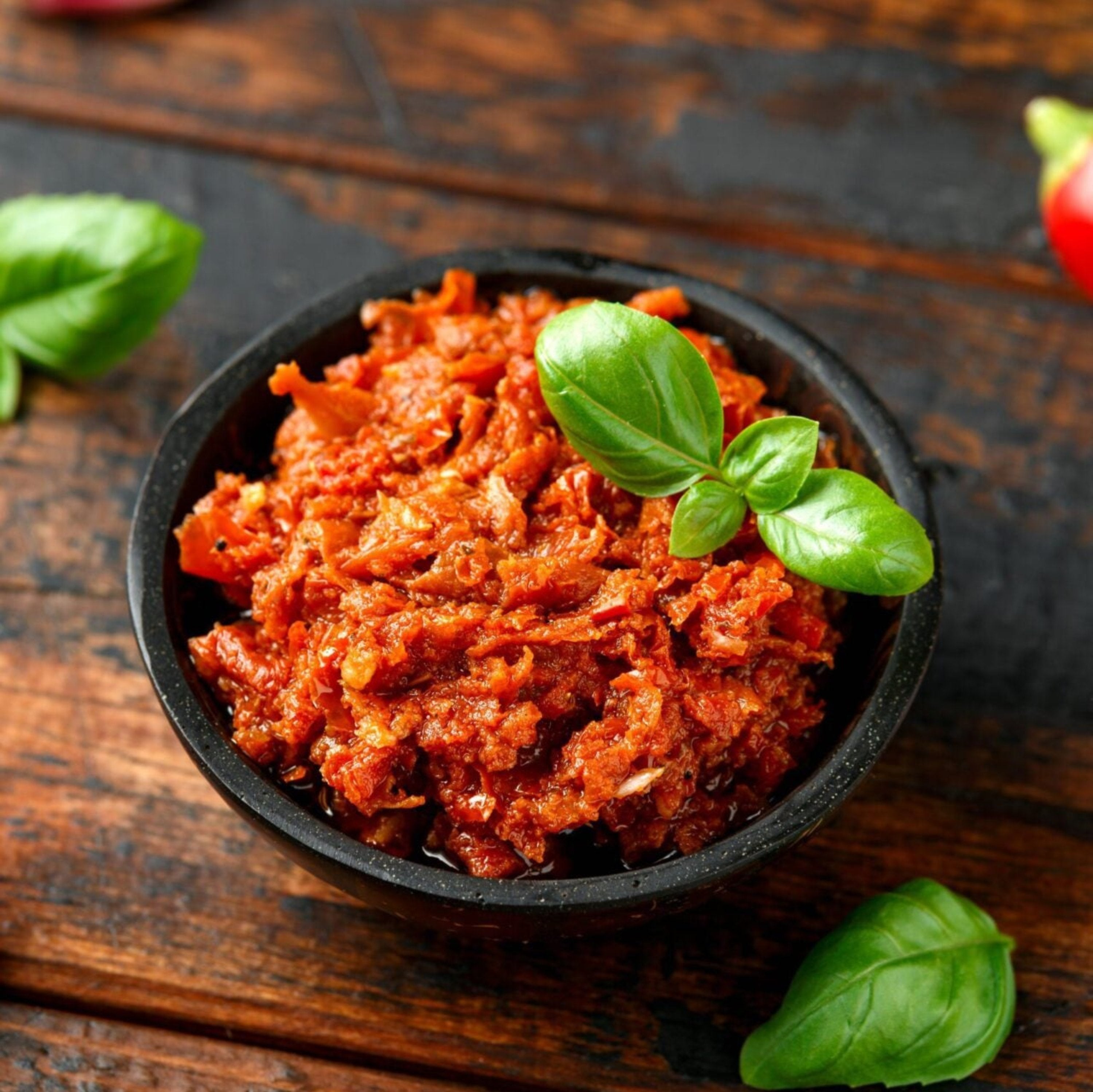 Rotes Knoblauch-Pesto in schwarzer Schale mit frischen Basilikumblättern auf rustikalem Holztisch