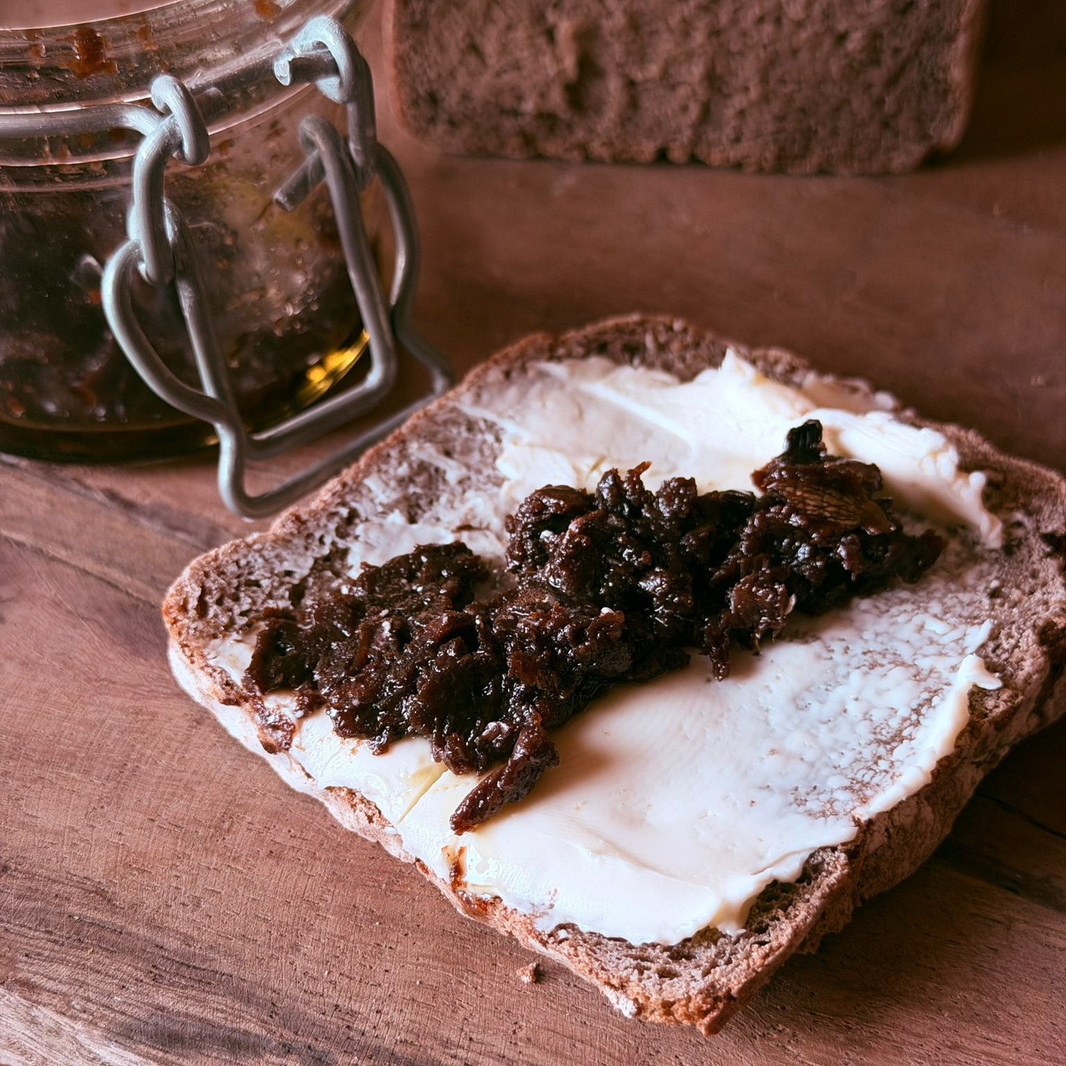 Brotscheibe mit Butter und dunklem Aufstrich aus Schwarzem Knoblauch mit Hanföl, dahinter ein Bügelglas