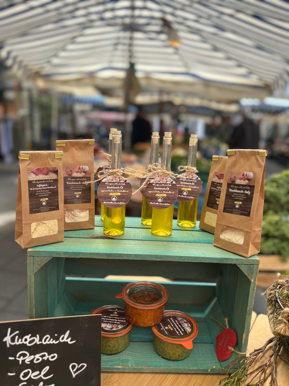 Marktstand der Knobi-Farm mit Gewürztüten, Knoblauch-Öl-Flaschen und Pesto-Gläsern auf einer türkisen Holzkiste